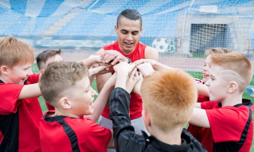 Portrait of junior football team stacking hands during motivational pep talk before match in outdoor stadium, focus on young coach, copy space