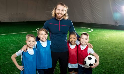 Successful football trainer and four happy boys in uniform standing on green soccer field